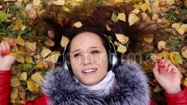 美丽幸福的年轻女子戴着耳机躺在秋叶上听音乐。 季节，科技和人